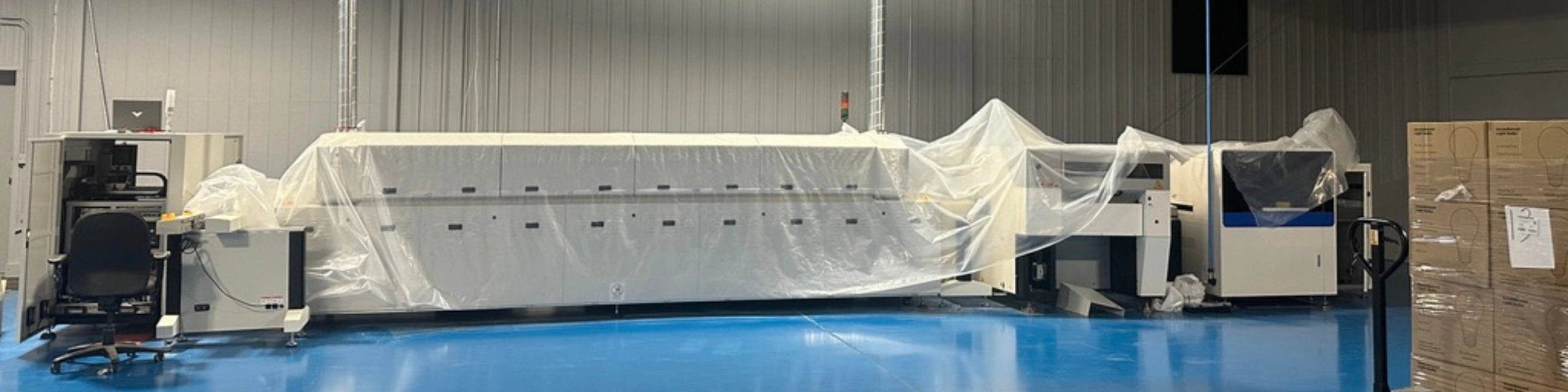 A large industrial bulb machine covered with a translucent plastic sheet, photographed inside a warehouse or factory with a blue floor and metal walls.
